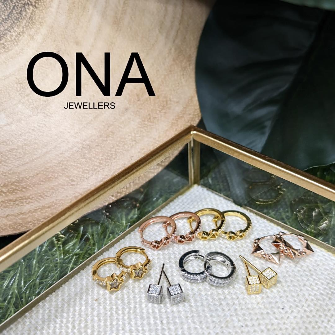 A display box with gold and silver rings and earrings, set on a glass surface with the text "ONA Jewellers" in the background.