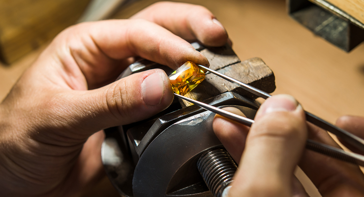 A person uses tweezers to set a yellow gemstone into a metal fixture, working with precision on a piece of jewelry.
