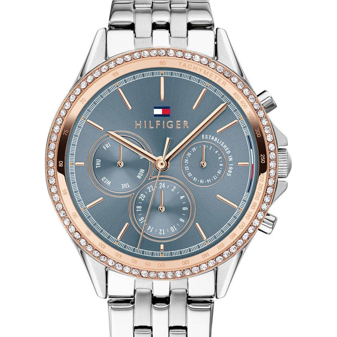The TOMMY HILFIGER 1781976 Ari is a blue and silver stainless steel ladies watch featuring a blue face, gold accents, three subdials, a crystal bezel, and a metal link band.