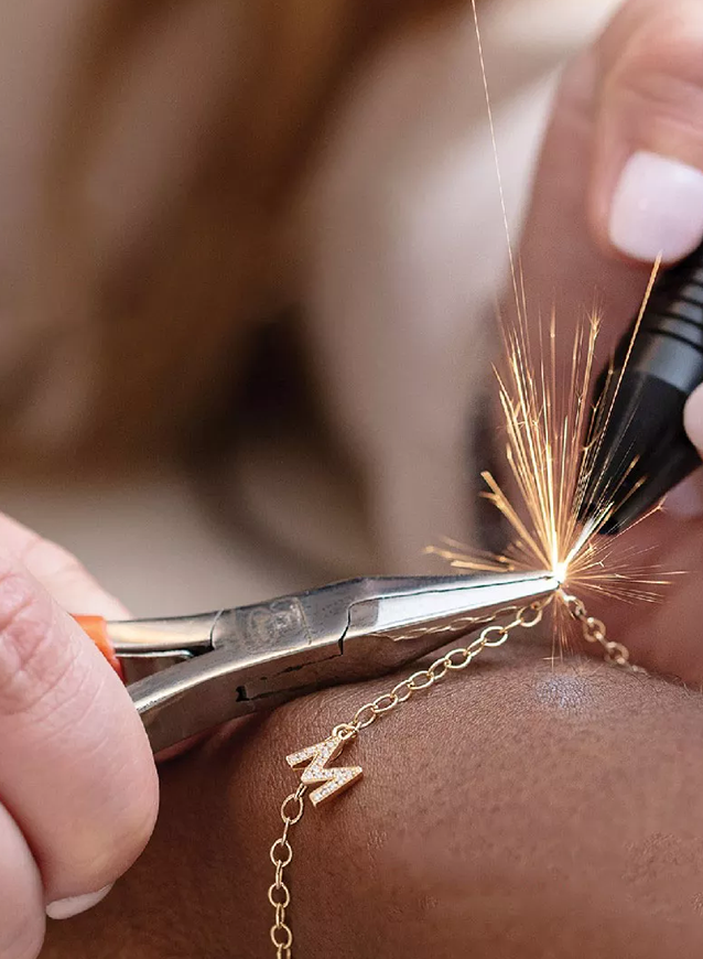 Close-up of hands using pliers and a tool to weld a gold chain bracelet with a small "M" charm, producing bright sparks.