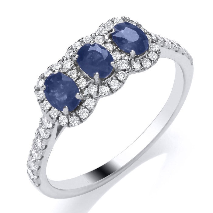This 9ct White Gold Sapphire and Diamond Trilogy Ring features three oval sapphires, each surrounded by sparkling diamonds, with additional diamonds set along the elegant shoulders.