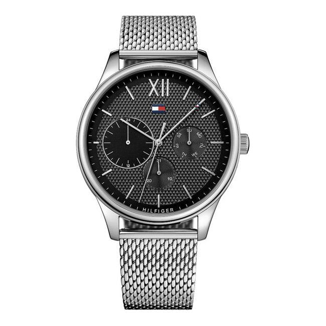 The Tommy Hilfiger 1791415 Damon Mesh Bracelet Watch features a silver mesh band, black textured dial, three subdials, silver hour markers, and the Tommy Hilfiger logo below 12 o’clock.