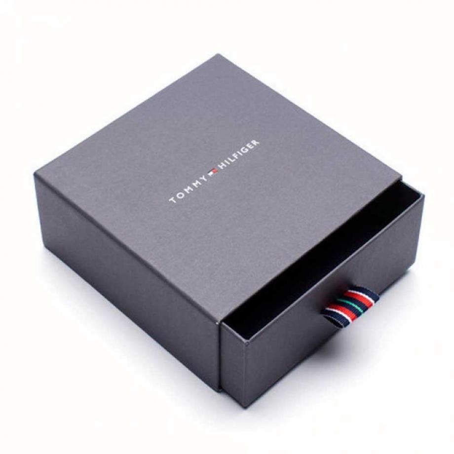 A partially opened Tommy Hilfiger gift box with a ribbon pull tab displays the logo on the lid and reveals the Tommy Hilfiger Stainless Steel Silver Tone Double Layer Coin Charm Necklace (2780067) inside.