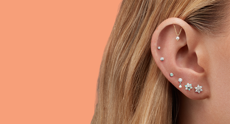 Close-up of a person's ear with multiple piercings, each adorned with different styles of small, silver and gold earrings, against a solid peach background.