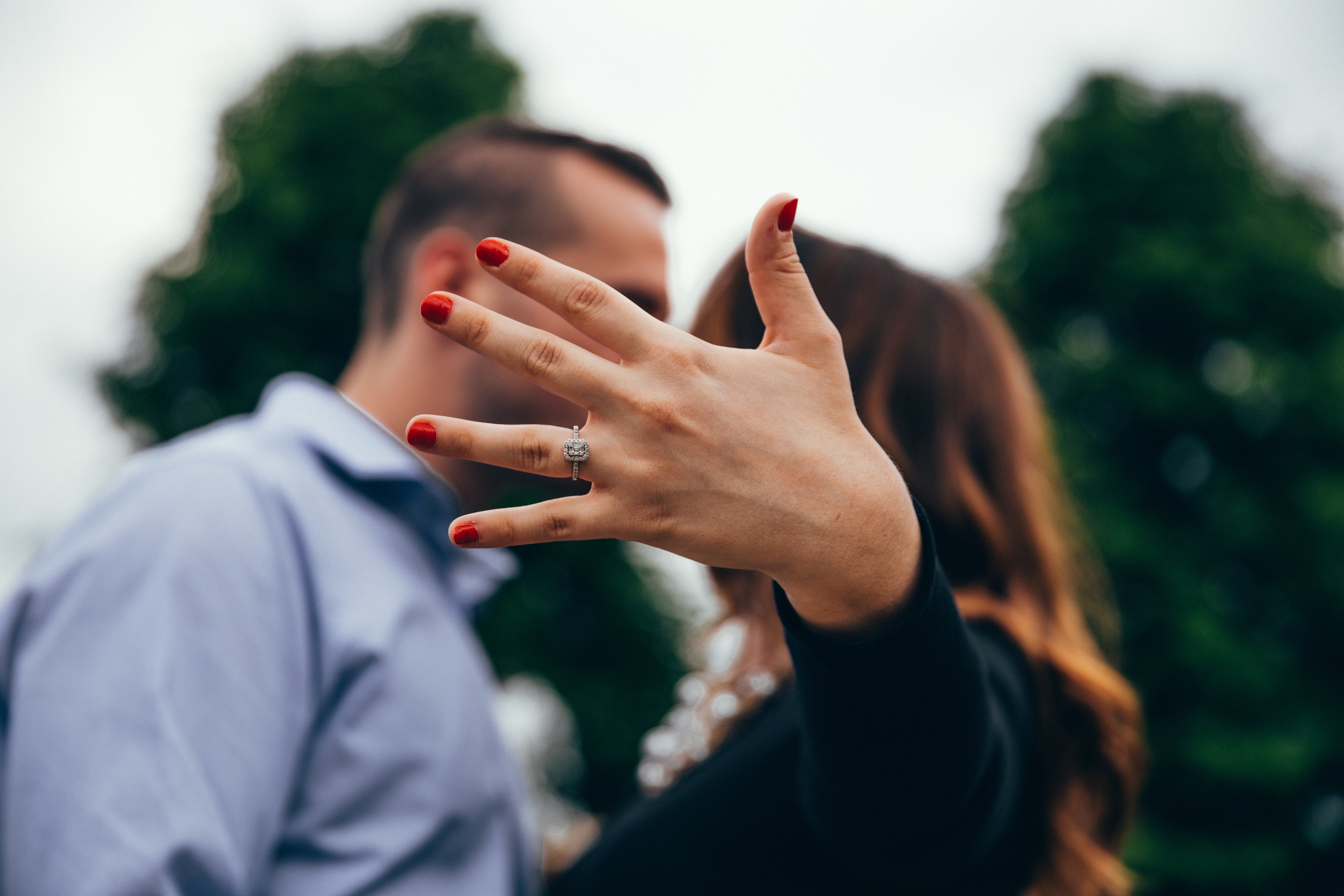 A person with red nail polish displays an engagement ring on their hand, with another person standing close by; both are outdoors with trees in the background.