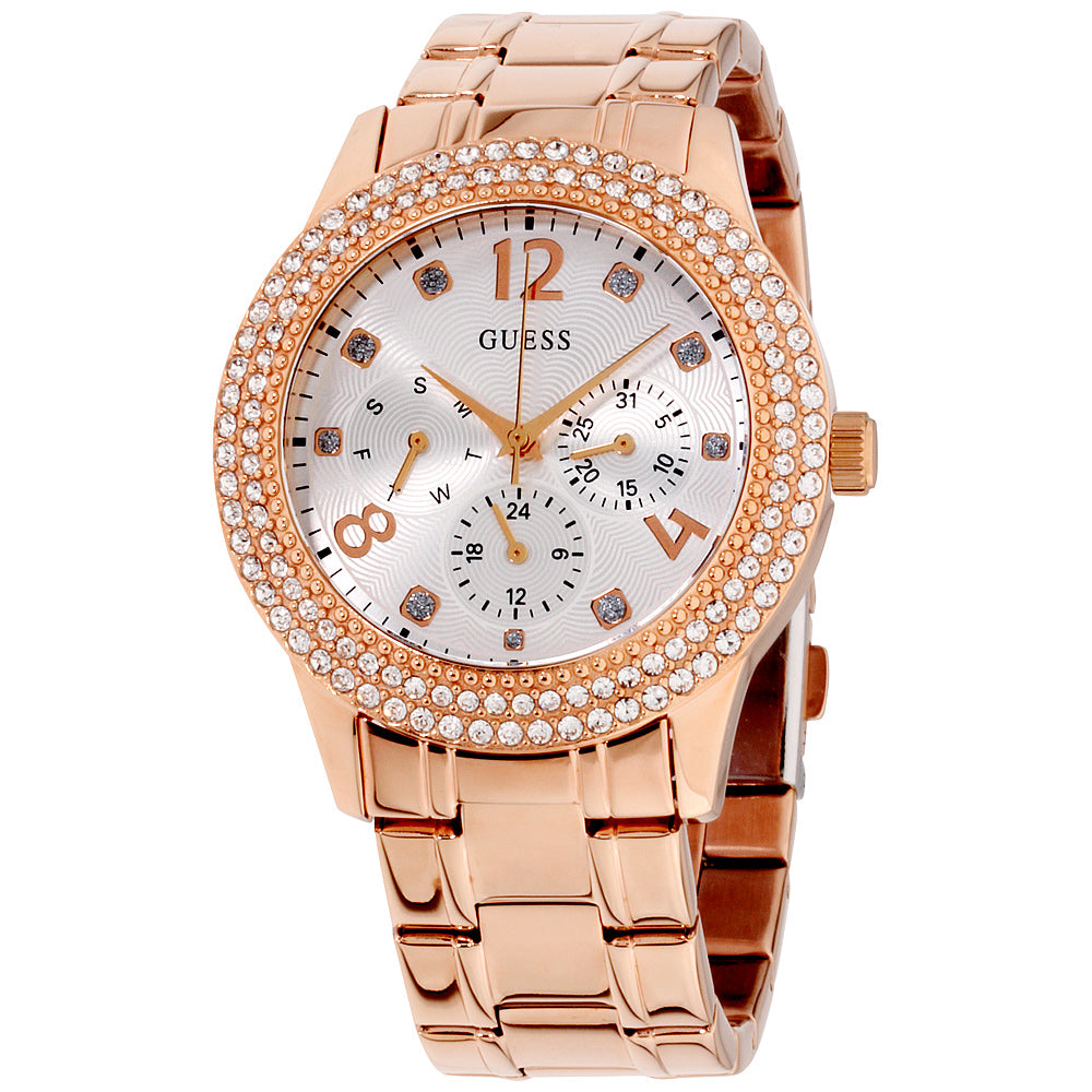 The Guess Ladies Bedazzle Watch W1097L3 features a rose gold-tone case, Swarovski crystal-studded bezel, silver dial with three subdials, and a stylish metal link bracelet.