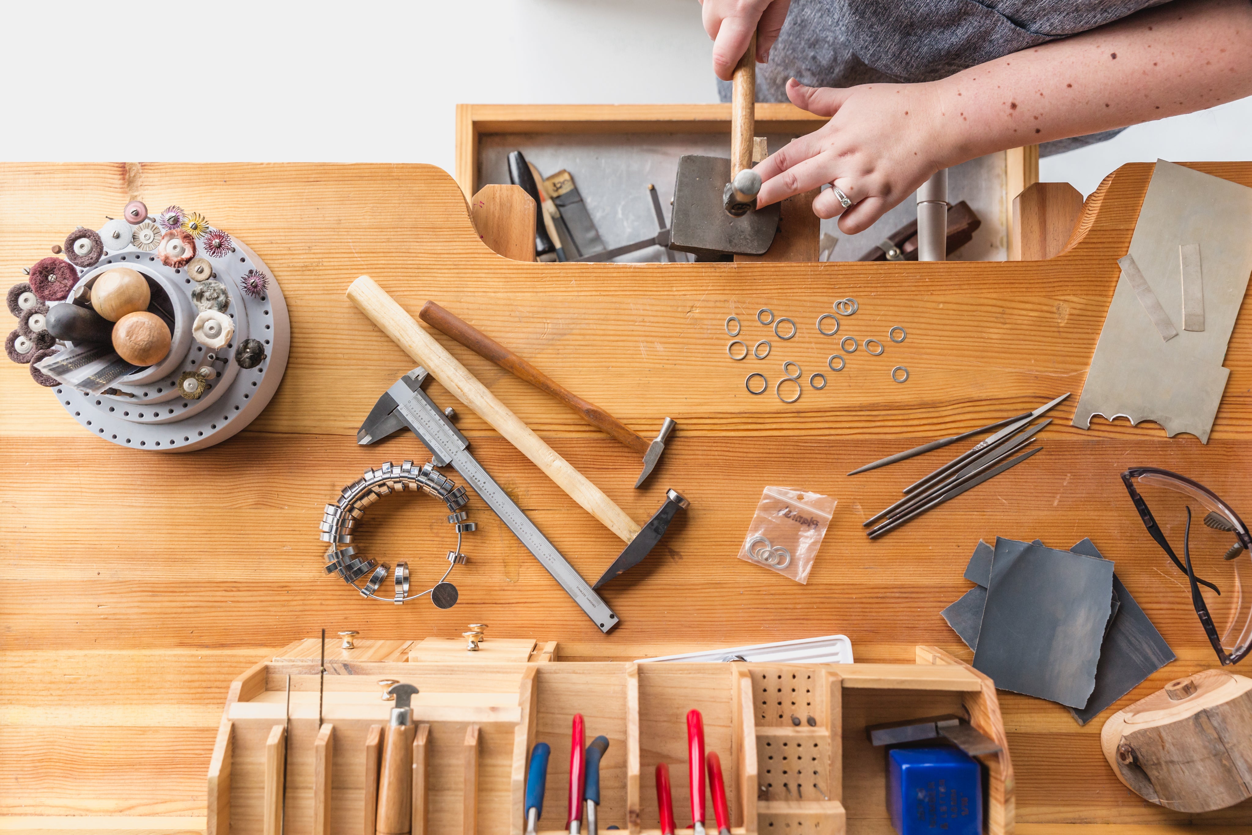 A person uses a hammer at a wooden workbench with various jewelry-making tools, metal rings, tweezers, and pliers organized in compartments.
