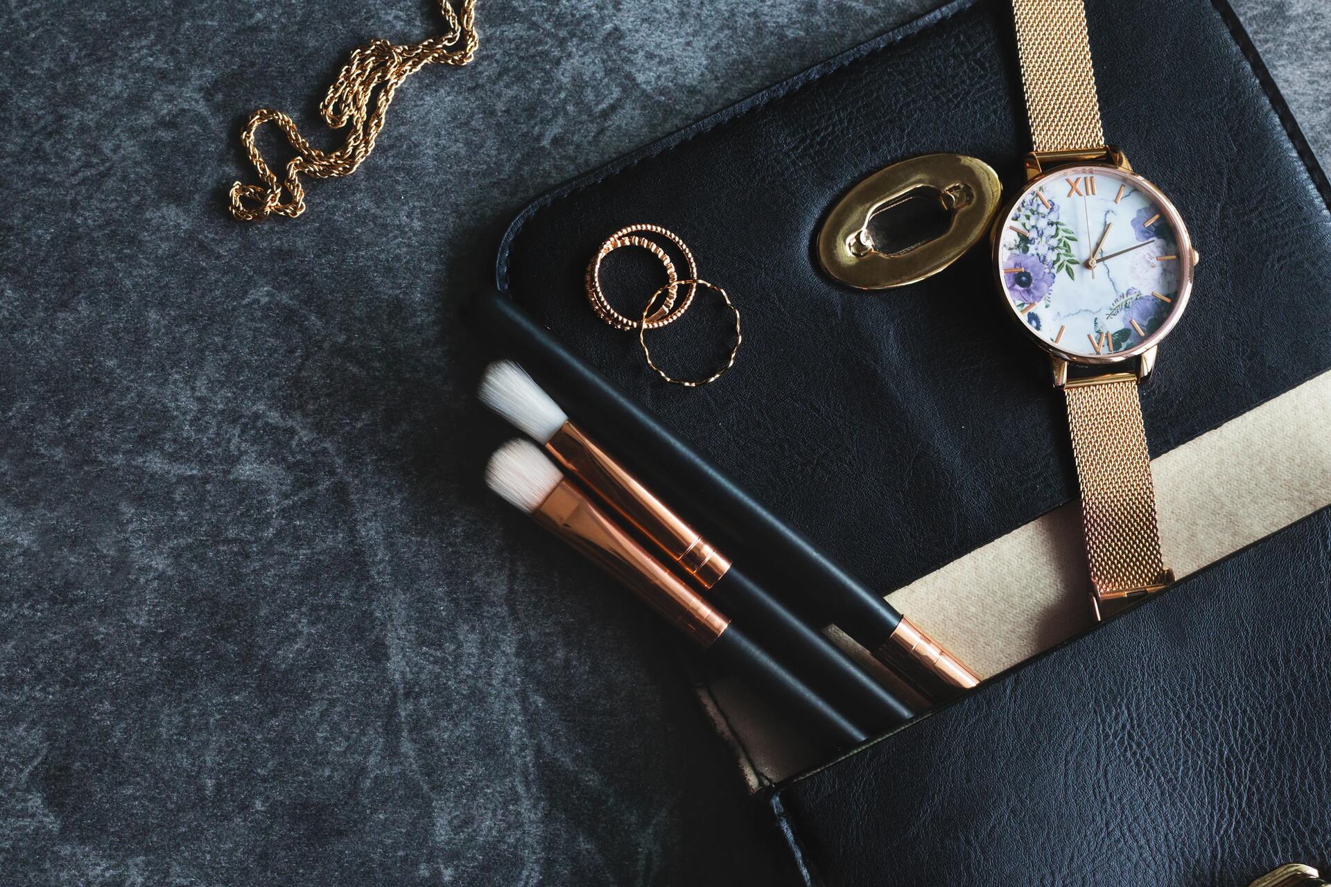 Rose gold watch, makeup brushes, and jewelry on a dark surface