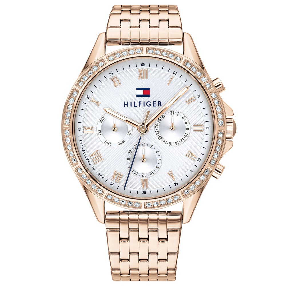 The Tommy Hilfiger Ari Women's Watch 1782143 features a gold-tone stainless steel strap, white dial with Roman numerals, three subdials, a crystal-studded bezel, and a linked metal band.